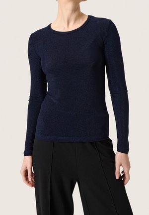 Vrouw die een aansluitend, langarmig marineblauw shirt draagt met een ronde halslijn en een zwarte broek met hoge taille tegen een effen achtergrond.