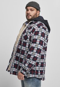 Camisa de franela a cuadros con patrones en azul marino, rojo, blanco y negro; forrada con forro polar beige; cuenta con botones y bolsillos en el pecho; llevada sobre un hoodie azul marino.