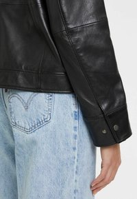Schwarze Lederjacke mit glatter Textur, schmal geschnitten und mit Druckknopffront verziert. Getragen über hellblauen Jeans mit herzförmiger Tasche.