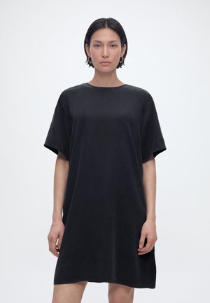 Femeie cu păr scurt și închis la culoare, purtând o rochie neagră lejeră cu mâneci scurte, stând în picioare pe un fundal simplu, gri deschis.