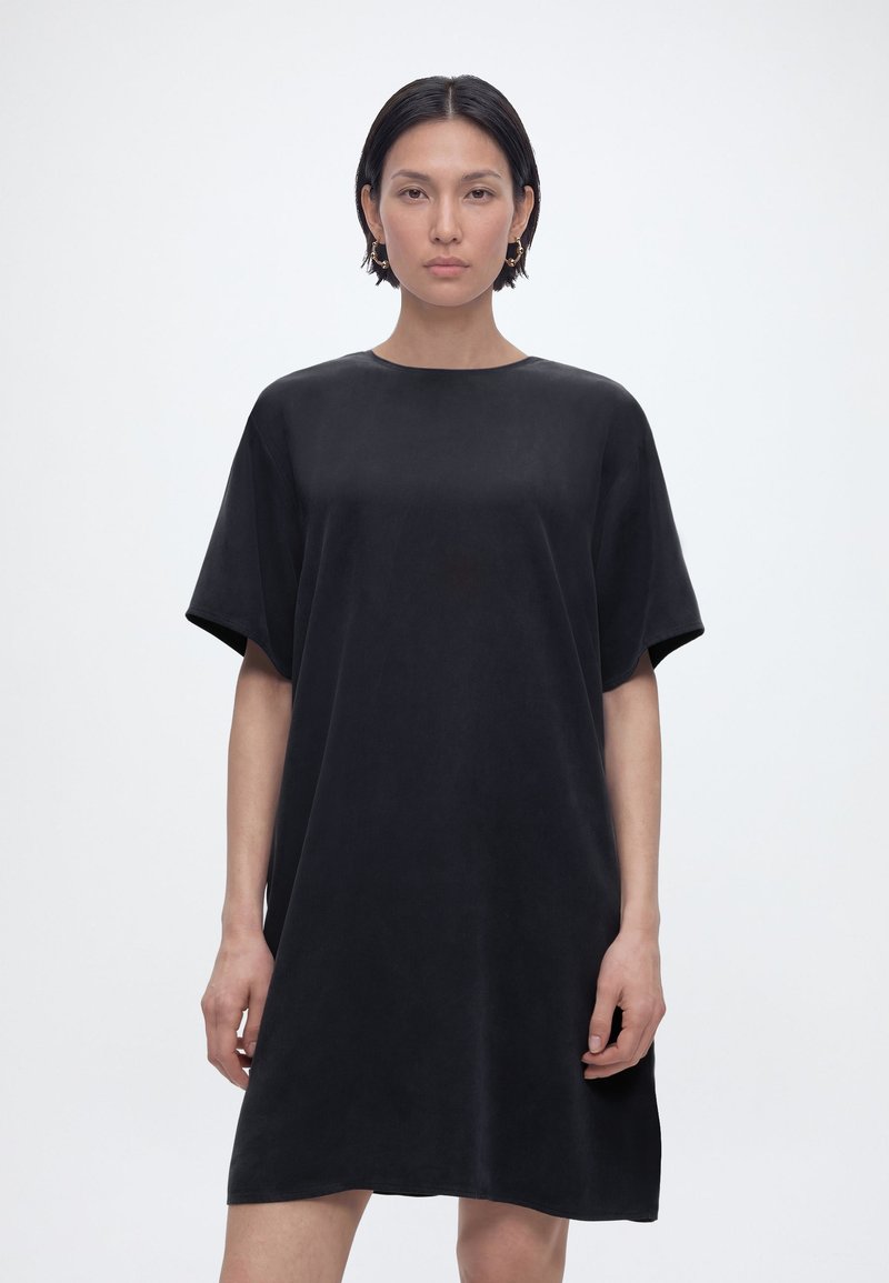 Γυναίκα με κοντά σκούρα μαλλιά που φοράει χαλαρό μαύρο φόρεμα με κοντά μανίκια, στέκεται μπροστά σε ένα απλό ανοιχτό γκρι φόντο.