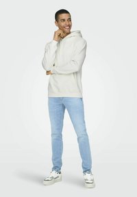 Hellbeige Hoodie mit einer Kängurutasche, verstellbaren Kordeln, kombiniert mit hellblauen Jeans und weißen Sneakers mit grauen Akzenten.