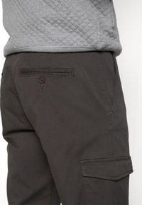 Gros plan d'un pantalon cargo foncé avec une poche arrière boutonnée et une poche latérale à rabat, porté avec un haut matelassé gris.