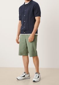 Marineblauw poloshirt met korte mouwen, gebreid en met gestructureerde strepen, knoopsluiting; gecombineerd met olijfgroene shorts en zwart/witte sportschoenen.