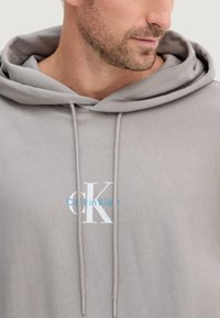 Felpa con cappuccio grigio chiaro con coulisse, caratterizzata da un grande logo CK bianco e "Calvin Klein" in azzurro chiaro. Il tessuto appare morbido e liscio.