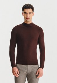 Brązowy sweter typu golf wykonan y z gładkiego dzianiny, dopasowany fason, z długimi rękawami i ściągaczem na dole, zestawiony z szarymi, fakturowanymi spodniami.