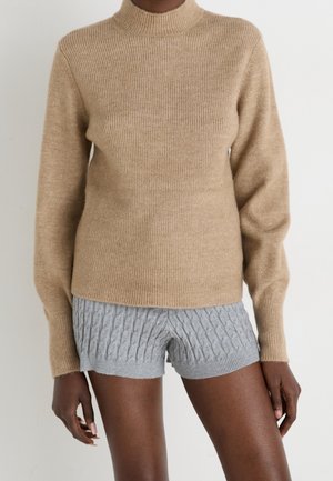 Personne portant un pull côtelé beige à manches longues et un short gris en maille torsadée sur un fond uni.