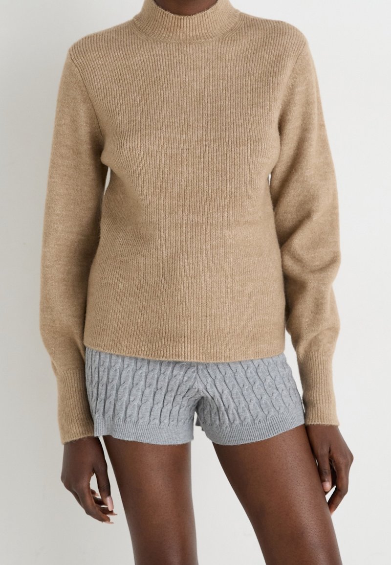 Personne portant un pull côtelé beige à manches longues et un short gris en maille torsadée sur un fond uni.