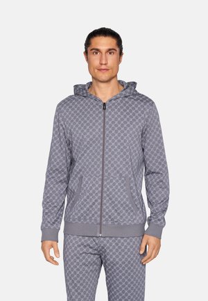 Mies, joka pukeutunut harmaaseen, kuvioituun vetoketjulla varustettuun huppariin ja housuihin, seisoo yksinkertaisen vaalean taustan edessä.