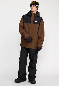 Veste imperméable marron et noire avec capuche, arborant un logo sur la poitrine. Associée à un pantalon noir et des gants, les chaussures sont noires.