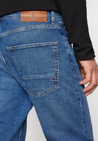 Modré džíny z denimu s voľným strihom. Medzi funkcie patrí prešité zadné vrecko a hnedá kožená nášivka značky na páse.