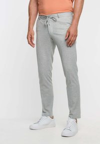 Man som bär ljusgrå stickade joggingbyxor med snörning, vita sneakers och en kortärmad persikofärgad tröja mot en vit bakgrund.