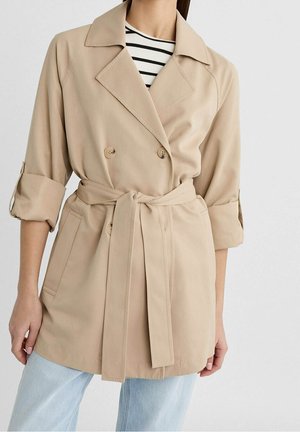 Femme portant un trench-coat beige ceinturé avec manches retroussées par-dessus un haut rayé noir et blanc et un jean bleu clair.