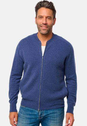 MarineblauesZip-Up-Cardigan aus weicherWolle mit runderHalsausschnitt sowie geripptenÄrmelbündchen und Saum. Getragen über einem weißen Hemd, kombiniert mit blauen Jeans.