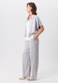 Hellgraue, kurz geschnittene Jacke mit Knopfleiste und elastischem Bund; kombiniert mit einer weit geschnittenen Hose aus passendem hellgrauem Stoff. Flache weiße Sandalen.