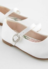 Ballerines blanches en tissu avec un bout rond, une sangle latérale ornée d'une boucle en strass ronde et une semelle intérieure à motif floral. Semelle en caoutchouc plate.