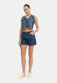 Completo in denim blu scuro composto da giacca corta e pantaloni shorts, con chiusura a bottoni, due tasche sul petto e orli arrotolati. Abbinato a scarpe da ginnastica chiare.