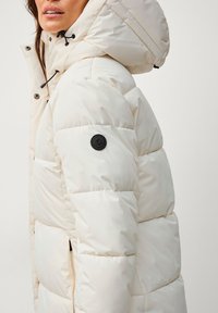 Doudoune couleur blanc cassé, dotée d'une capuche, de pièces en métal noir et d'un patch logo sur la manche gauche ; texture matelassée et design mi-long.
