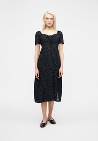 Γυναίκα που στέκεται φορώντας μαύρο midi φόρεμα με λευκές πουά, μανίκια με όγκο, τετράγωνο ντεκολτέ και καφέ επίπεδα παπούτσια σε απλό φόντο.