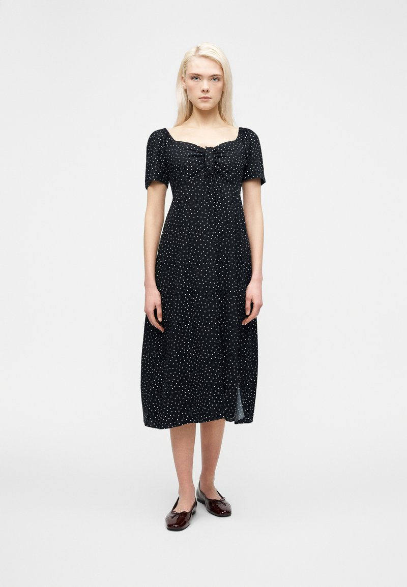 Γυναίκα που στέκεται φορώντας μαύρο midi φόρεμα με λευκές πουά, μανίκια με όγκο, τετράγωνο ντεκολτέ και καφέ επίπεδα παπούτσια σε απλό φόντο.