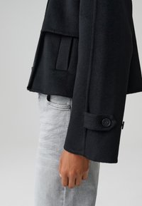 Giacca in pile nera con colletto alto e polsini con bottoni, caratterizzata da una tasca laterale e un design strutturato, abbinata a jeans grigi.