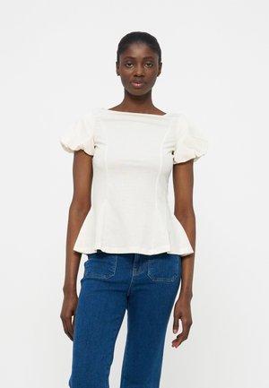 Kobieta o ciemnej karnacji i włosach związanych do tyłu, ubrana w biały top z bufkami na rękawach i niebieskie dżinsy, na tle jednolitej białej ściany.