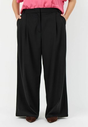 Persona che indossa pantaloni neri ampi e larghi con tasche laterali, abbinati a una camicetta rosa e scarpe marroni, in piedi con le mani nelle tasche.