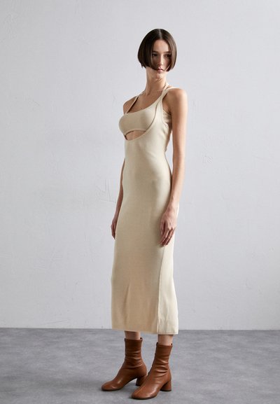 Beige knit dress with cut-out detail, slim fit, and spaghetti straps, paired with brown ankle boots. Smooth texture, mid-length design.