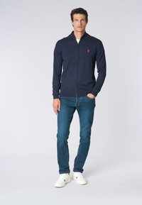 Maglione zip-up blu navy con colletto alto, polsini e orlo a costine, caratterizzato da un piccolo logo rosso sul lato sinistro del petto, abbinato a jeans blu e sneakers bianche.