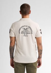 Beżowa koszulka polo z czarnymi grafikami na plecach, z wizerunkiem orła i napisem. Krótkie rękawy z łatami na ramionach, materiał z teksturowanej bawełny.