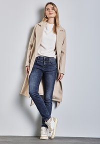 Manteau long beige sur un pull blanc, associé à un jean bleu et des baskets beiges. Tissu lisse, coupe droite et détails minimaux.