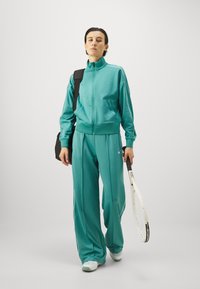 Zelenomodri trenirka z visokim ovratnikom, zadrgo spredaj in kontrastnimi belimi stranskimi črtami, v kombinaciji s črno torbo in teniško loparjem.