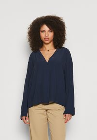 Blusa a maniche lunghe color blu navy con scollatura a V, vestibilità rilassata e maniche ampie. Tessuto morbido, dettagli minimi, abbinata a pantaloni khaki.