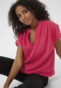 Blouse à manches courtes et décolleté en V en tissu rose vif, avec une texture douce et des fronces aux épaules. Associée à un pantalon noir.