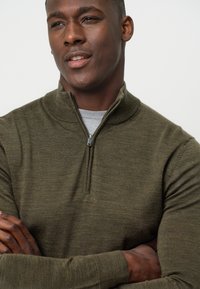 Olivgrüner Reißverschluss-Sweater mit einem Stehkragen, gerippten Manschetten und strukturiertem Strickdesign. Das Model steht mit verschränkten Armen, darunter trägt es ein graues Hemd.