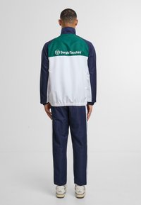 Giacca da allenamento bianca e blu scuro con dettagli verde, caratterizzata da un colletto alto e orlo elastico. Logo visibile sul retro. Indossata con pantaloni blu scuro abbinati.