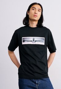 Sort bomulds T-shirt med et frontgrafik af en basketballspiller. Korte ærmer, rund halsudskæring, afslappet pasform og flad kant.