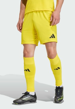 Mann trägt gelbe Sportshorts, passende Socken mit schwarzem Logo und schwarze Stollenschuhe mit neonfarbenen Akzenten vor hellem Hintergrund.