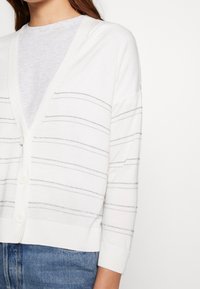 Vit cardigan med silverfärgade horisontella ränder, långa ärmar, V-ringning och knappstängning. Mjukt, lätt material, avslappnad passform.