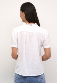 Hvid linned kortærmet skjorte med en flæsekrave, der har en samlet ryg og en let buet kant. Båret med blå jeans.