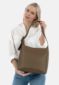 Donna in camicia bianca e jeans blu che tiene una grande borsa in pelle marrone per il manico su uno sfondo semplice.