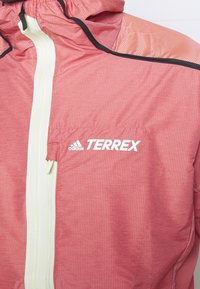 Rote, leichte Jacke mit Kapuze, die weiße Reißverschlüsse und das "TERREX"-Logo aufweist. Strukturiertes, knitterartiges Muster und schwarze Akzente an den Rändern.