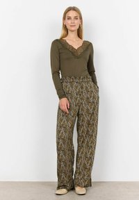 Langærmet oliven-grøn bluse med blonde detaljer ved halsen, parret med mønstrede, løstsiddende bukser i jordfarver og beige flade sko.