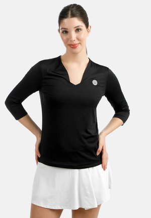 Mujer con camisa negra de manga larga y cuello en V con un pequeño logo y falda blanca plisada, posando con las manos en las caderas.