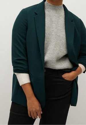 Personne portant un blazer vert foncé sur un pull en tricot gris clair, avec un pantalon noir, une main dans la poche et l'autre détendue sur le côté.