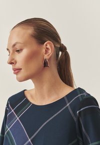 Tropfenohrringe in Maroon mit glänzendem Finish, eingerahmt von goldfarbenem Zubehör. Das Modell trägt ein marineblaues Oberteil mit karierten Akzenten.