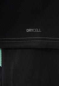 Musta urheilu- ja treenikangas, jossa on teksturoitu pinta. Kankaassa on ommeltu sauma ja harmaa "DRYCELL" -logo teal-värisen sivustuksen yläpuolella.
