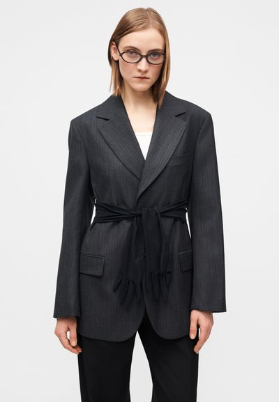 Grå blazer med smalle striber og brede revers, med sort bælte bundet i taljen og klaplommer, kombineret med en hvid top.