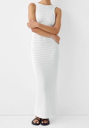 Mouwloze witte maxi-jurk van gehaakt katoen, gedragen met zwarte platte sandalen, met de armen gekruist over de taille tegen een effen witte achtergrond.