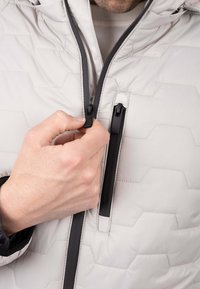 Chaqueta gris claro con un patrón acolchado hexagonal, que cuenta con una cremallera negra y detalles en negro. Se muestra una mano cerrando la cremallera de la chaqueta.